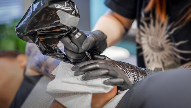 Tattoo artist at work. Visible hands in black gloves holding tattoo gun with needle and drawing a line on a customer upper arm.