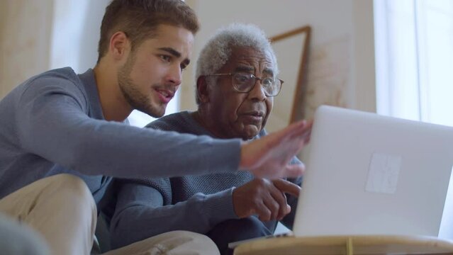 Bearded Caucasian guy helping senior Black man with using laptop. Young man sitting on couch with his grandpa while explaining things and pointing at screen. Family bonding, generations concept.