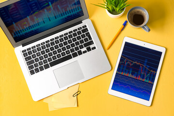 Laptop and tablet computer with stock data and cup of coffee on yellow background. Finance trading
