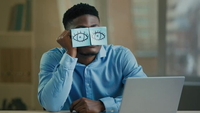 Sleepy lazy office worker businessman african american ethnic man sleeper sit at desk put on face funny comical stickers fake eyes exhausted tired weary employee napping fall asleep relaxing sleeping