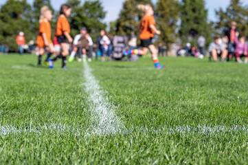 Selective focus on ground level view of soccer field center line with defocused youth girls in...