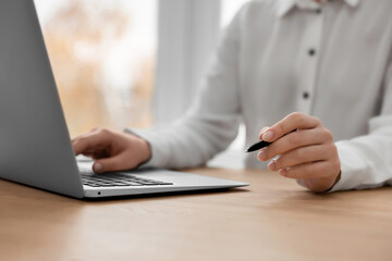 Woman with pen working on laptop at wooden table, closeup. Electronic document management