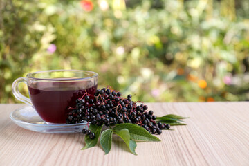 Glass cup of tasty elderberry tea and Sambucus berries on wooden table outdoors, space for text