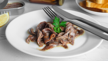 Plate with canned anchovy fillets, fork and knife on white table, closeup