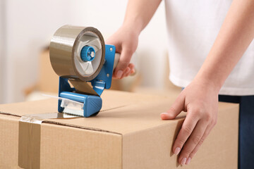 Woman applying adhesive tape on box with dispenser indoors, closeup