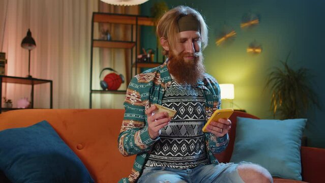 Bearded hippie man using credit bank card and smartphone while transferring money, purchases online shopping, order food delivery at home apartment indoors. Young redhead guy in evening room on couch