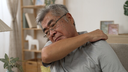 frowning elderly asian man is enduring awful ache on shoulder and massage by hand while sitting on couch in living room. close up view of illness old male suffering neck pain on sofa. health concept