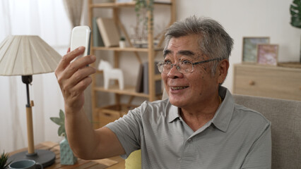 grandfather make video call to relatives with smartphone at home. Concept technology modern generation family connection. old asian Japanese man waving hand on couch and talking online by cellphone
