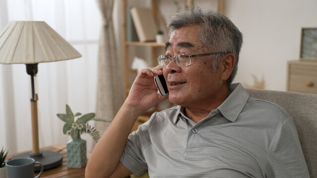 Smiling asian old man talking on smartphone on sofa. Mature male in online conversation using mobile phone while laughing. cheerful elderly guy in glasses having fun during funny conversation call