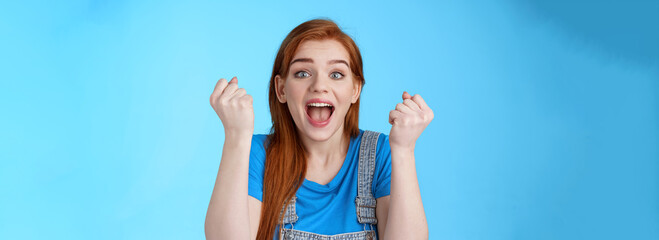 Naklejka na ściany i meble Close-up cheerful lucky redhead woman win lottery, fist pump yell yeah hooray, winning bet, smiling gladly rejoicing excellent news, celebrating success, triumphing happily, stand blue background