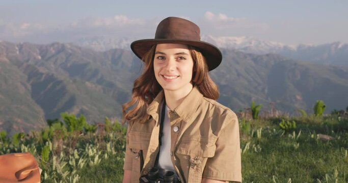 Female environmentalist smiling and laughing as she rests in middle of patrol in the mountains, sitting in the grass. Woman security guard in the mountains with binoculars looking at camera, smiling.