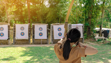 Back view of Asian girl wear face mask aims archery bow and arrow to colorful target in shooting...