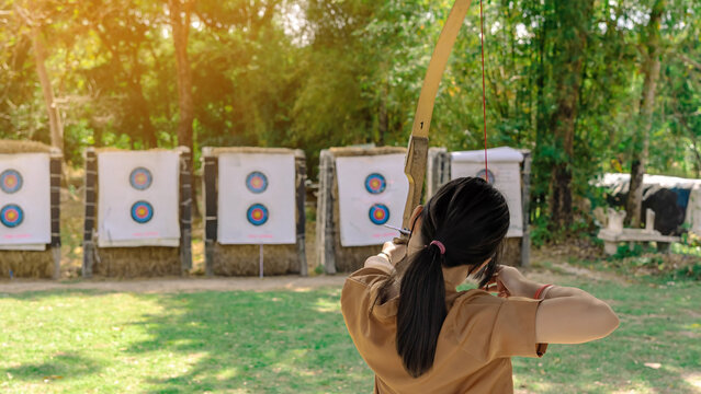 Back view of Asian girl wear face mask aims archery bow and arrow to colorful target in shooting range during training and competition. Exercise and concentration with outdoor archery.Archery for teen