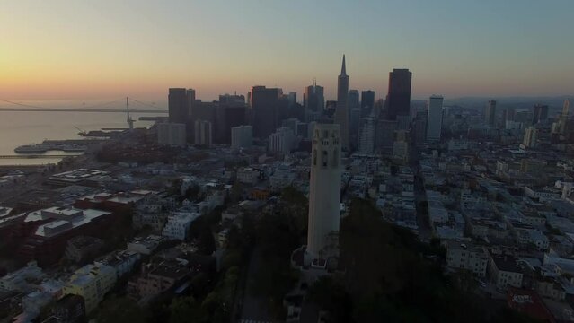 Drone shot pulling away from Coit Tower standing amid San Francisco's downtown skyline at sunset.