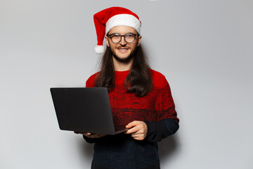 Studio portrait of handsome smiling guy, holding laptop in hands. Wearing Santa Claus hat and eyeglasses. Christmas concept. White background.