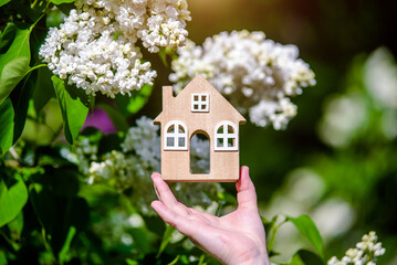 The girl holds the house symbol against the background of blossoming white lilac
