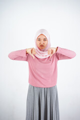 Beautiful Asian woman in pink sweater and hijab shows upset expression with thumbs down on white background