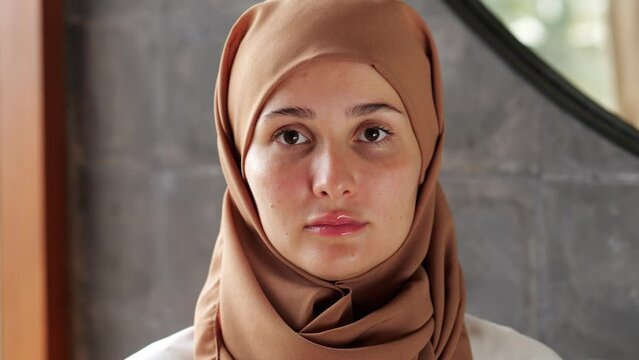 Young woman in a hijab, sitting with her head slightly tilted forward and her eyes down, close-up portrait. She calmly raises her head and looks confidently straight into camera
