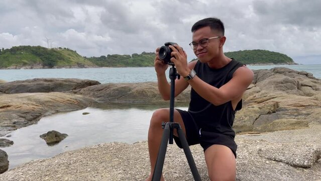 Filipino young man on his knees testing dslr camera focus on tripod and ocean behind. Asian photographer taking photos in natural landscape in Phuket, Thailand. Tourism, travel destination concepts