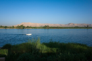 Nile river landscape in Luxor, Upper Egypt, White motorboat on blue water with view of Sahara desert dry mountains and green plantations on river west bank