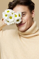 Obraz na płótnie Canvas Close-up photo.A beauteous young man with fair skin, with dark, short, beautiful hair combed back, in a beige turtleneck with a high collar stands on a dark beige background with a bouquet of daisies