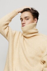 A handsome young man with dark short hair combed back in a beige sweater with a high neck stands on a gray background with a hand on his head .Vertical studio shot.