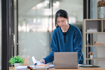 Fototapeta premium Young Asian businesswoman standing at her office desk and working on her laptop with happiness.