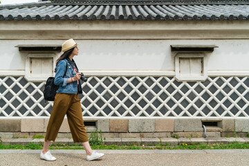 full length with side view Asian female photographer passing by Japanese traditional building with diamond pattern wall while visiting Osaka castle in japan on sunny day © PR Image Factory