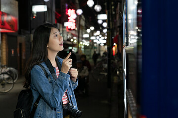 hesitant korean girl standing in front of vending machine thinking about which drink to choose on shopping street in Osaka japan at night. she looks at items with finger pointing