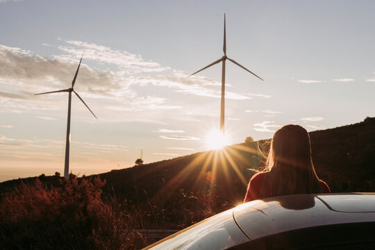 Woman Leaning On Car Looking At Wind Park At Sunset