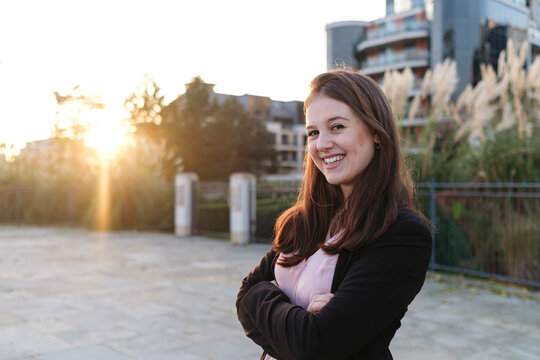 Happy young woman with arms crossed on footpath at sunset