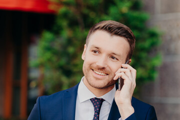 Headshot of handsome unshaven young male has conversation with service operator, has happy expression, attractive look, dressed in formal outfit, poses outdoor. People and lifestyle concept.