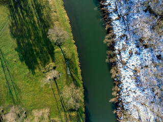 A birds eye view of the winter river Skawa near Wadowice, Poland. Surrounded by forest, with snow on the grass. Shadows of the trees. Aerial view.