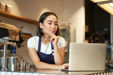 Portrait of smiling korean woman, barista in coffee shop, standing at counter with laptop, smiling and looking confident, self-employed female entrepreneur in her own coffee shop - obrazy, fototapety, plakaty
