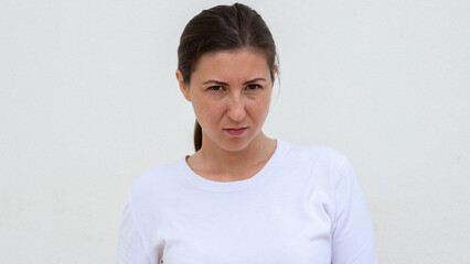 Portrait of grumpy young woman looking at camera over white background. Caucasian woman wearing white T-shirt expressing disgust or irritation. Anger concept