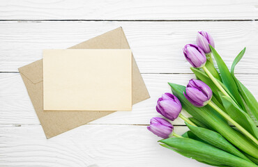 Bouquet of purple tulips and blank greeting card on white wooden background, top view, copy space.