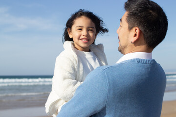 Cropped image of beaming father and daughter on beach. Japanese family walking, hugging, dad carrying little girl. Leisure, family time, parenting concept