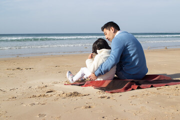 Happy Japanese family on blanket on beach. Father and daughter in casual clothes sitting on blanket, hugging. Family, relaxation, nature concept