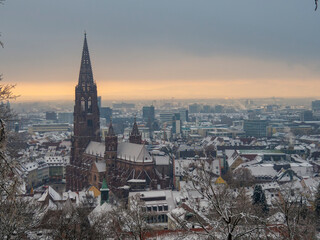 Obraz premium View of the snow-covered Freiburg Minster Cathedral against sunset sky during winter. Snowy skyline of old town panorama. High viewpoint with winter landscape from the Black Forest hill.