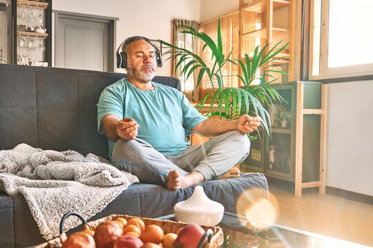 Mature middle-aged overweight man in wireless headphones relaxing at home with guided meditation, listening to relaxing music on smartphone and meditating in lotus pose.