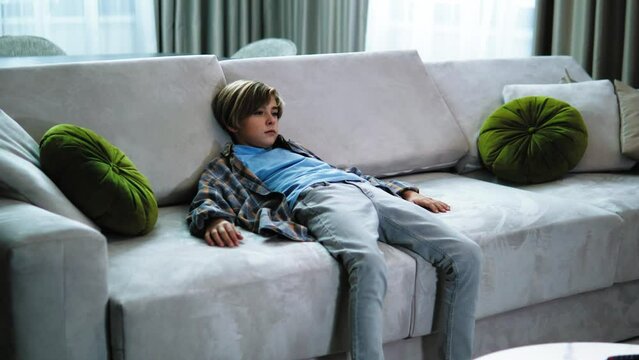 Tired exhausted handsome boy teenager walking and then falling on the sofa in the living room at home. Boy is relaxed on the coach.  Child kid boy sitting on sofa, chilling, doing nothing, so tired 
