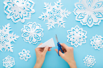 Young adult woman hands cutting different white snowflake shapes from paper on light blue table background. Pastel color. Making decoration elements for winter festive. Closeup. Top down view.