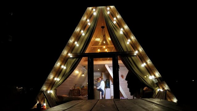 Mother and son are having fun and dancing in a one-story wooden bungalow house with lanterns in the evening and a triangular roof descending to the ground. Winter vacation in the forest.