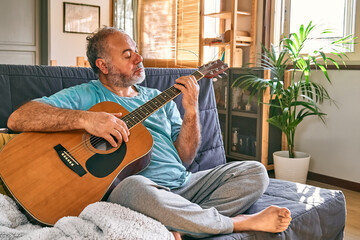 Middle-aged bearded man playing acoustic guitar while sitting on sofa in light living room.