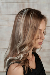 Close-up shot of a blonde girl with ash blonde wavy hair extensions. The girl in a black top with clip-in fake hair strands looks down on a light wooden background. Side view.