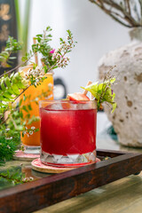 Close-up glass of raspberry juice placed on a wooden tray with colorful flowers and leaves decorated around. Blurred foreground and background, selective focus