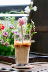 Close-up of a glass coffee cup placed on a wooden table with colorful flowers and leaves decorated around. Blurred foreground and background, selective focus