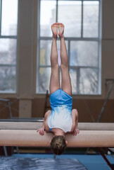 A young girl gymnast performs a handstand on a balance beam in a gym