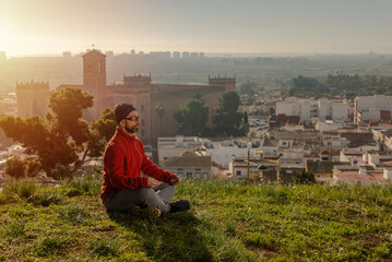 Man sitting cross-legged, meditating, on a hill with landscape of a small European town at sunrise