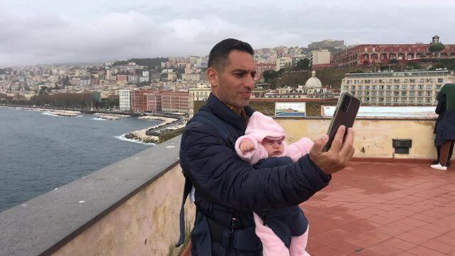 Father with small toddler girl in carrier baby sling. Travel with child. Man make picture selfie on observation deck overlooking sea and city, talking by video on phone. outdoor